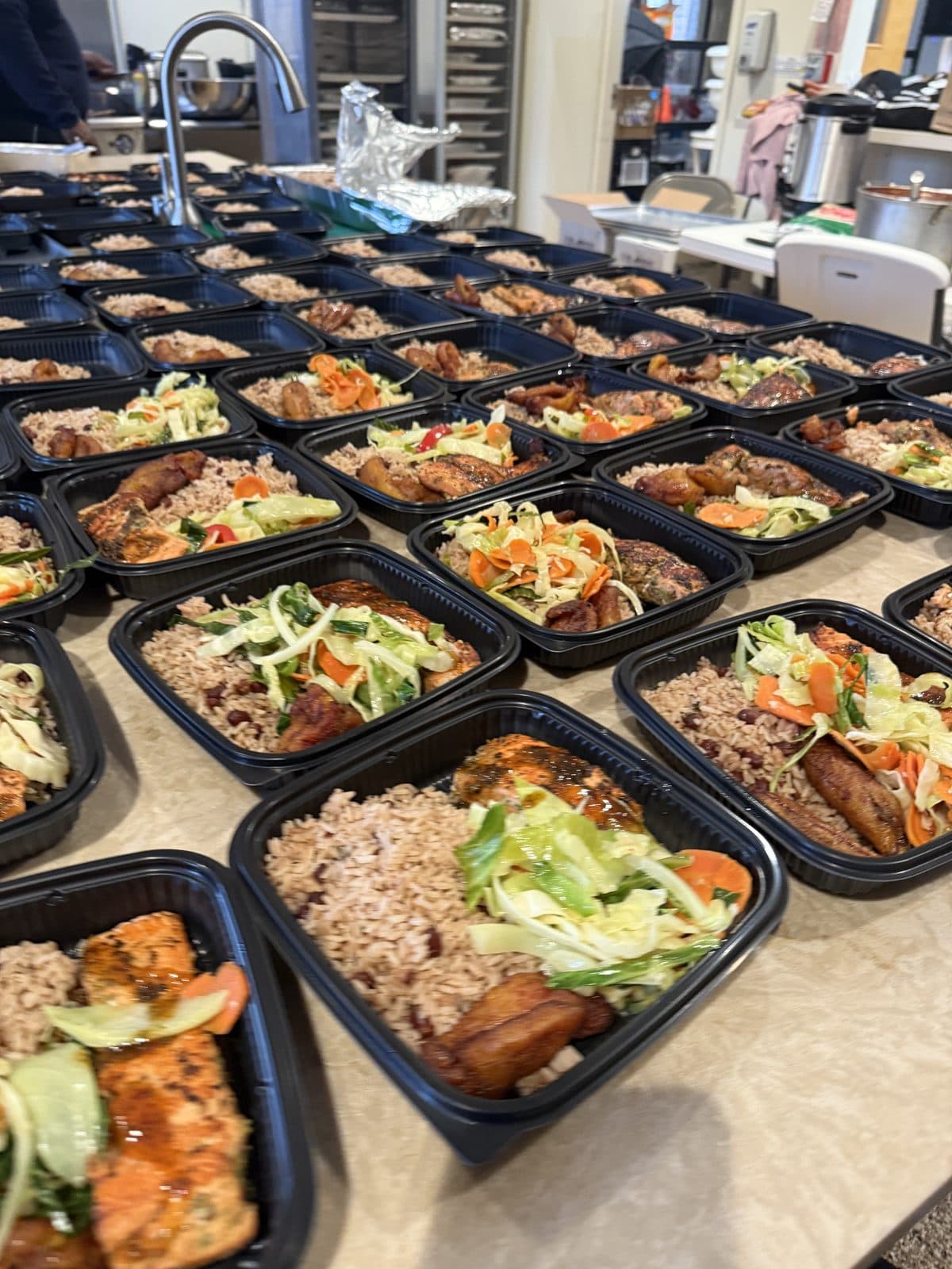 Rows of packed meal-prep containers with jerk chicken, rice and peas, slaw, and plantain